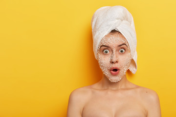 Daily morning routine concept. Embarrassed young woman stands shirtless, applies peeling facial mask for removing dark dotes on face, stares at camera wears white soft towel on head isolated on yellow