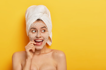 Photo of glad European girl with toothy smile, uses sea salt for spa procedures, takes shower, has smooth healthy skin, looks away, wears white towel, isolated over yellow background. Beauty concept