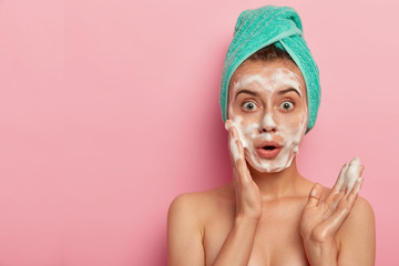 Photo of surprised European woman washes face with foam gel, wants to have refreshed well cared...