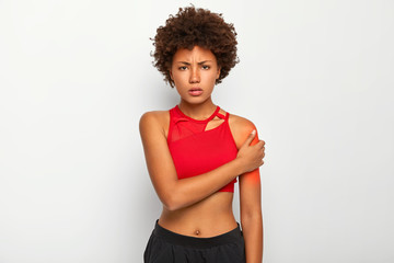 Photo of displeased Afro American woman with slim figure, dressed in red top and black trousers, touches shoulder, suffers from pain, being injured, poses against white background. Painful feeling