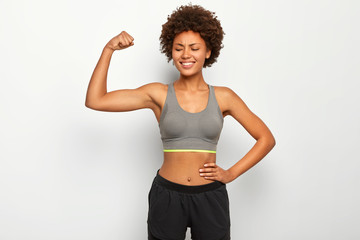Obraz na płótnie Canvas People, sport and strength concept. Glad curly haired African American woman raises arm, shows biceps, demonstrates muscles, has slim figure, wears casual top and shorts, models over white wall