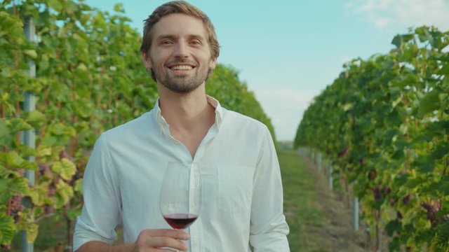 Portrait of positive smiling handsome young man in white suit tasting wine holding wineglass enjoying vacation at vineyard.