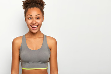 Fototapeta na wymiar Horizontal shot of happy curly woman has bright smile, shows white teeth, dressed in casual sport bra, has fitness or workout during spare time, models indoor. Healthy lifestyle, sport and emotions