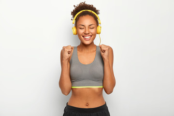 Plakat Studio shot of happy African American woman raises clenched fists, celebrates successful won game, dressed in sports bra, listens music via headphones, has slim body curly hair. Fitness female relaxes