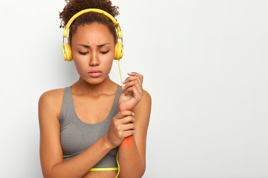 Young woman has injured wrist, touches arm with red pain location, has sad expression, listens favourite music with yellow stereo headphones, wears grey top, isolated over white studio wall.