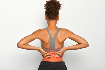Fototapeta na wymiar Indoor shot of faceless curly woman touches problematic zone of back, suffers from painful cramps, dressed in casual active wear, isolated over white background. Health care and medical concept