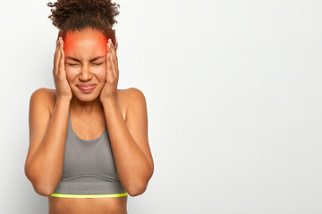 Fototapeta na wymiar Horizontal shot of dissatisfied curly haired woman touches both temples, suffers from migraine, has marked red location of pain, wears sports bra, closes eyes from bad feelings, isolated on white wall