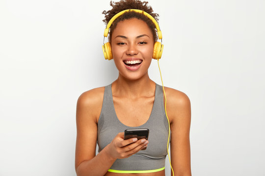 Sporty woman in active wear, smiles broadly, uses modern cell phone, listens music via headphones, chooses favourite track, poses against white background. People, active lifestyle and workout
