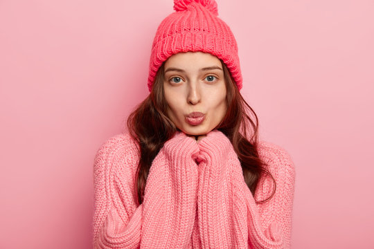 Portrait of beautiful young European woman keeps lips rounded, hands under chin, looks directly at camera, wears warm winter hat and sweater, poses over pink background. Cold weather concept