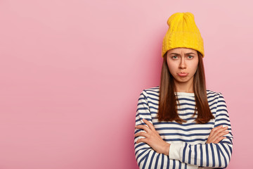 Pretty offended woman looks with sorrowful expression, frowns face and purses lips with displeasure, keeps arms folded, wears stylish yellow hat and striped sweater, models over pink wall, empty space
