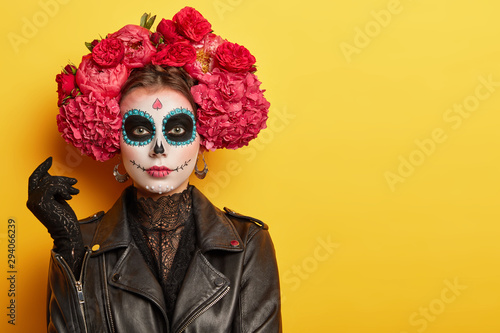 Photo of young woman has face arfully painted to resemble skulls, wears black leather jacket and gloves, wears garland made of red aromatic flowers, supports spiritual journey of dead people