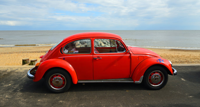 Classic Red Beetle parked on seafront promenade with sea and beach in background.