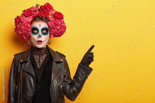 Surprised young woman has image of spooky ghost, has clay skull face, professional makeup wears red garland made of odorous flowers points away with scared expression invites going in mysterious place