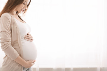 Happy pregnant woman touching her belly near window - obrazy, fototapety, plakaty