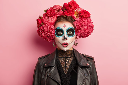 Shocked terrified young woman has scary ghost face, wears artistic makeup for Day of Dead holiday, wears black leather jacket, models over rosy studio background. Skull female symbolizing death
