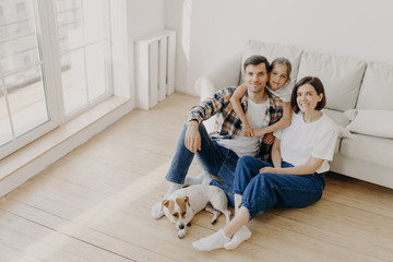 Lovely European family of father, mother and their daughter sit on floor near sofa in spacious...