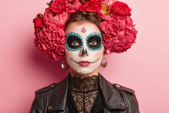 Portrait of calm female celebrates Day of Death, has sugar skull makeup, dark circles near eyes, painted smile, thinks death is natural part of human cycle, wears traditional mexican attire.