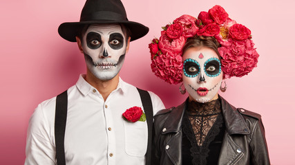 Surprised zombie man and spooky female wear mexican makeup, celebrate day of dead, wear black hat, white shirt traditional flower wreath ready to go on cemetery gather to pray and remember dead people