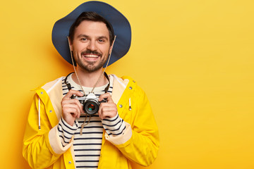 Positive handsome man holds retro camera, makes wonderful photos during trip, enjoys recreation time, smiles pleasantly, dressed in active wear, poses against yellow background. Active lifestyle