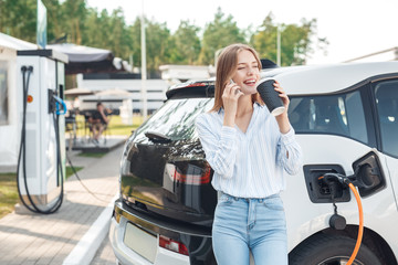 Young adult woman drinking coffee, charging electric car, talking on smartphone - obrazy, fototapety, plakaty