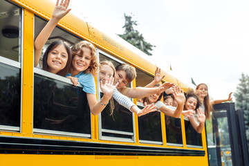 Classmates going to school by bus leaning out of the window waving to camera smiling positive