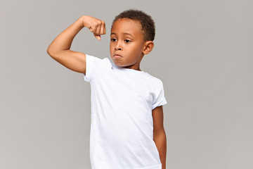 Cute dark skinned athletic male child with short Afro hairdo tensing arm muscle being proud of...
