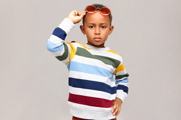 Kids fashion, style, childrens wear and accessories concept. Serious confident African American boy modeling against blank studio wall background wearing striped jumper and pink shades on his head