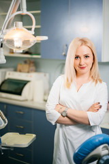 Fototapeta premium Professional female dentist in white doctor coat in dental chair and equipment, looking at camera and smiling. Confident woman posing during working process