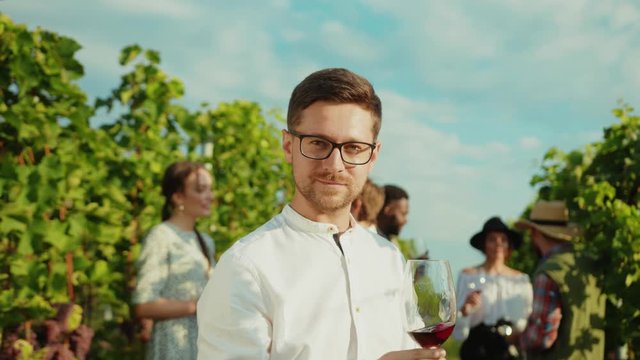 Attractive intelligent man in white shirt watching red color of tasty wine enjoying private tour holiday with friends in background. Vineyard.