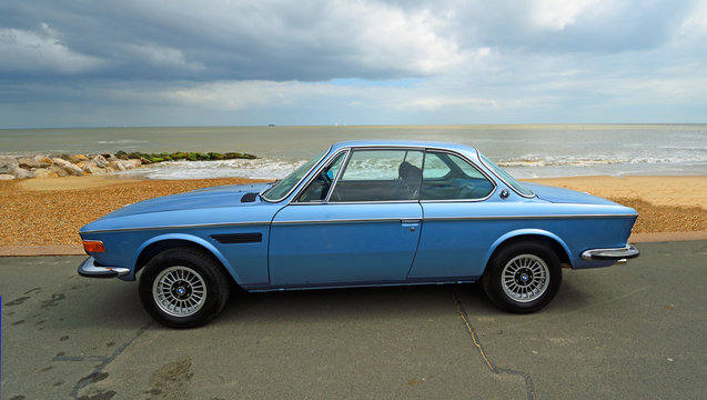  Classic Blue BMW Motor Car parked on seafront promenade with beach and sea in background.