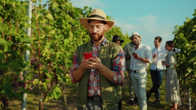 Handsome happy farmer in hat using application on smartphone in behind young luxury people tasting grapes and wine on vineyard.