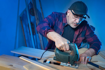 Sawing wood by electric jigsaw. The carpenter sawed wood detail. Work in the carpenter's shop. A man with an electric jigsaw on a blue background. Sale of woodworking tools.
