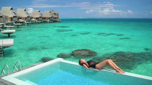 Luxury hotel Tahiti vacation swimsuit model woman relaxing sunbathing by infinity pool at overwater bungalow resort villa in Tahiti, French Polynesia. Suntan sexy Asian beauty on ocean beach travel.