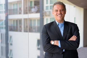 Happy mature Hispanic businessman smiling with arms crossed by the glass window of office building