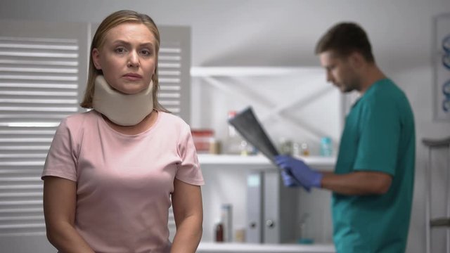 Sad female in foam cervical collar looking at cam, doctor examining x-ray result