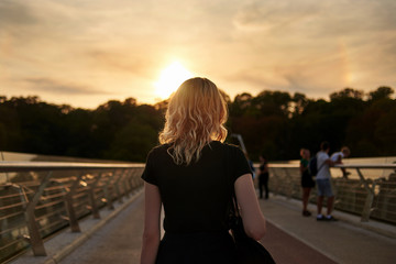 A young woman walks over a city bridge at sunset, back view.