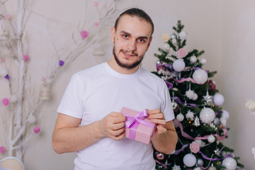 The guy with the beard in a white T-shirt holds a pink box. Bearded man with a gift in his hands in the new year. Christmas decor