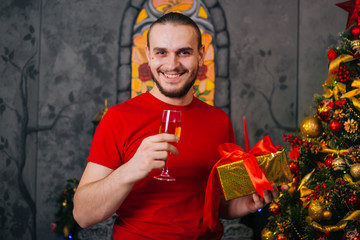 A man with a beard in a red T-shirt with a gift in hand and a glass of champagne. Festive mood, preparing for the new year. Christmas interior