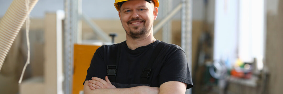 Portrait of young attractive man in work clothes and yellow helmet crossed arms smiling at shop for manufacturing furniture and details home interior