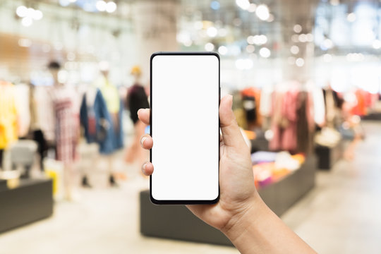 Mockup mobile phone image of woman hand holding smart phone with blank white screen for your advertisement on blurred abstract background of women clothing fashion shop.