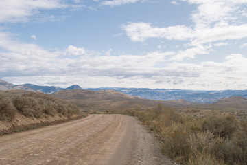 Fototapeta na wymiar Dirt road in the rural part of Kamloops, BC, Canada. Lac Du Bois Grasslands, vast open space with fields, hills, forested mountains, desert like landscape