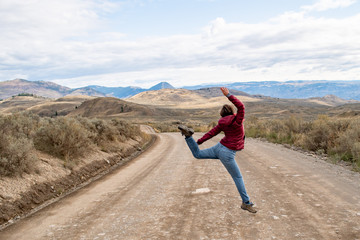 Young caucasian girl woman running and jumping ahead with determination the dirt road in rural BC, dry grass covered hills. Enjoying the freedom off road trip adventure