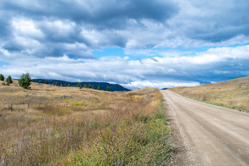 Fototapeta na wymiar Winding dirt road in the rural British Columbia near Kamloops. dry grassland, bush and shrub, occasional conifers trees, hills, desert like landscape. Road less traveled