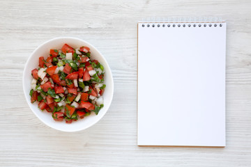 Pico de Gallo in a white bowl, blank notepad on a white wooden surface, top view. Overhead, from above, flat lay. Copy space.