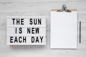'The sun is new each day' words on a lightbox, clipboard with blank sheet of paper on a white wooden surface, top view. Overhead, from above, flat lay. Copy space.