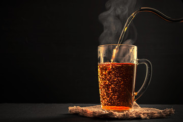 The process of brewing tea, pouring hot water from the kettle into the Cup, steam coming out of the mug, water droplets on the glass, black background