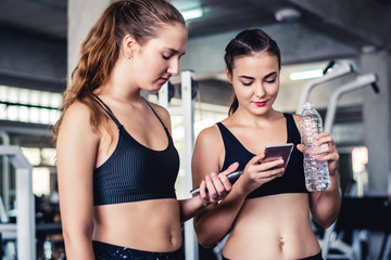 Fototapeta na wymiar Two young attractive Caucasian woman talking together and using smart phone or cellphone while drinking water and relaxation after hard workout at gym