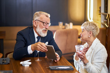 Attractive senior couple use modern technologies, tablet in restaurant. Grey-haired man in tuxedo, glasses and woman in white blazer