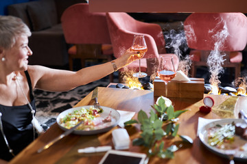 Mature elegant woman drinking champagne in restaurant. dishes with salad, ring, tablet, e-cigarette on table. Restaurant covered by light smoke from fireplace. Modern and trendy