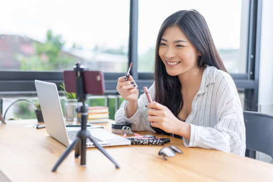 Young Asian cute artist woman beauty vlogger or blogger doing a cosmetic makeup tutorial vlog with brushes looking camera smartphone,recording viral clip share on social media live streaming Internet